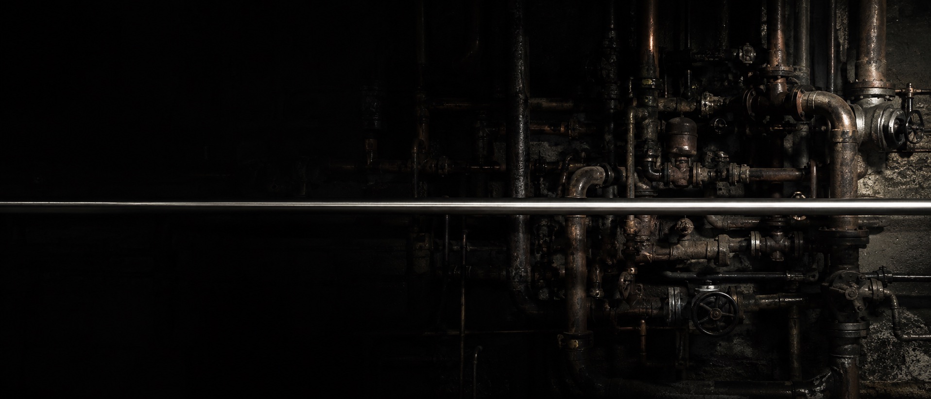 A wall of tangled industrial pipework with one straight polished pipe running through it